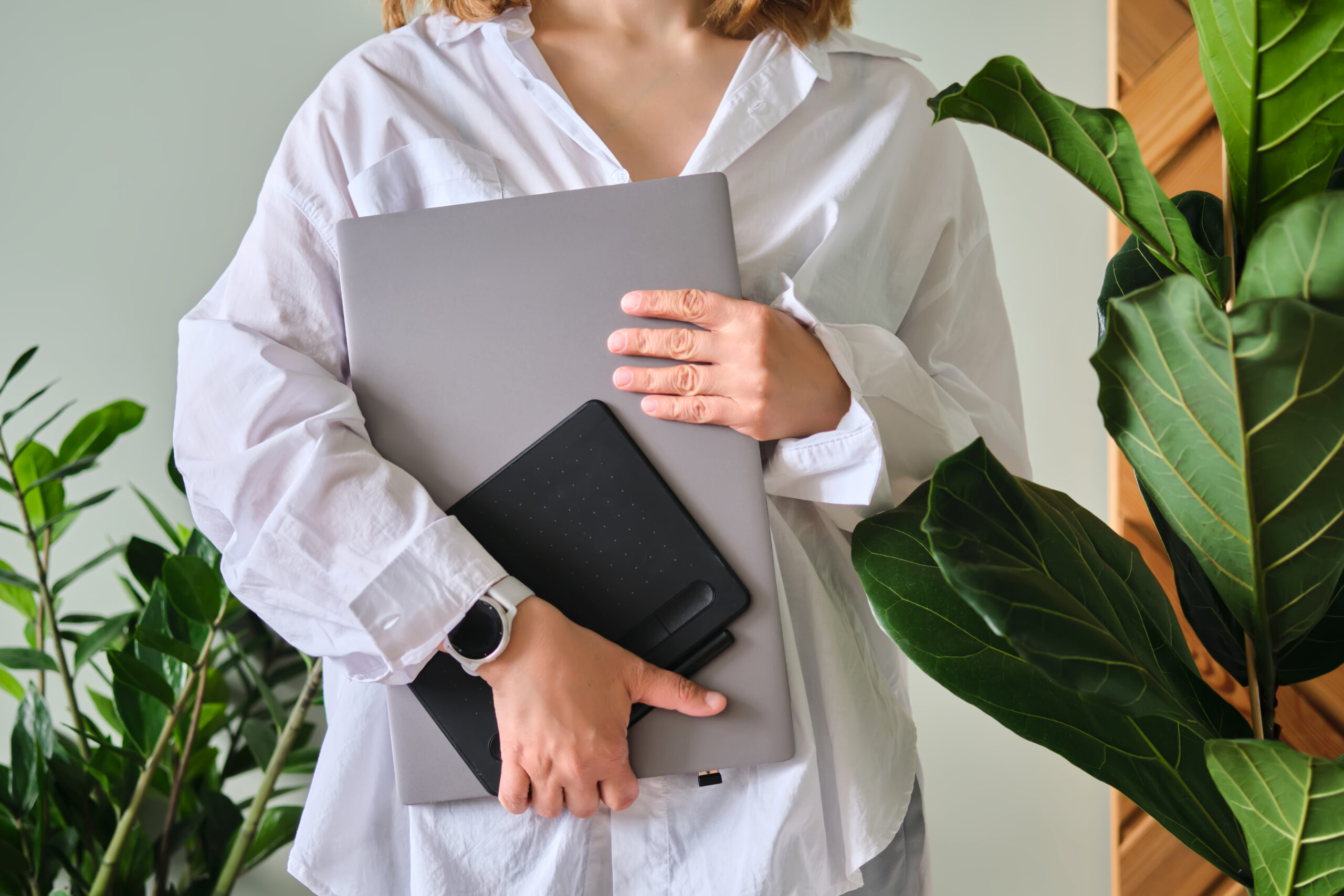 A woman holds a laptop and graphics tablet against a background of greenery.