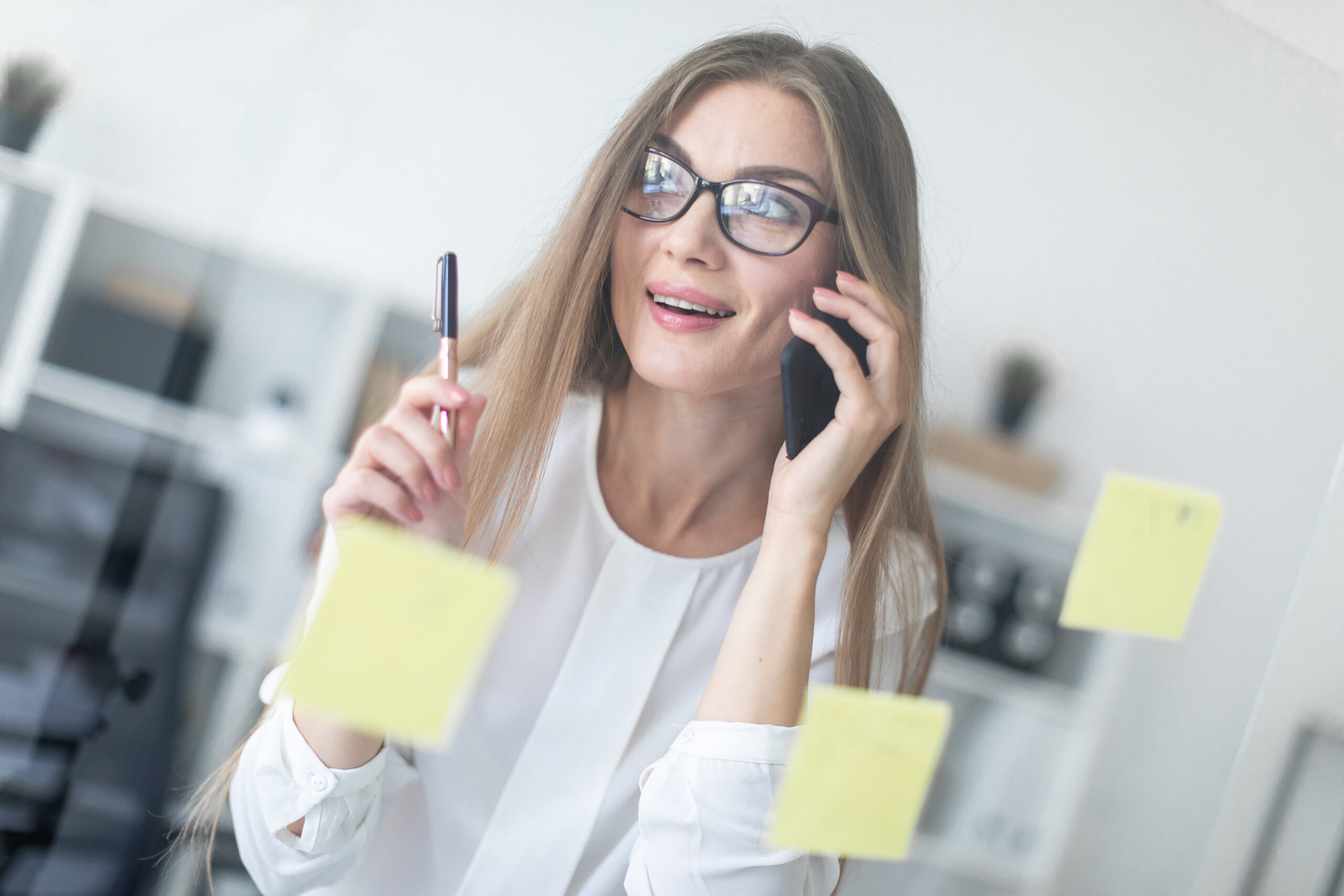 A young girl stands near a transparent board with stickers and holds a pen in her hand and talks on the phone.